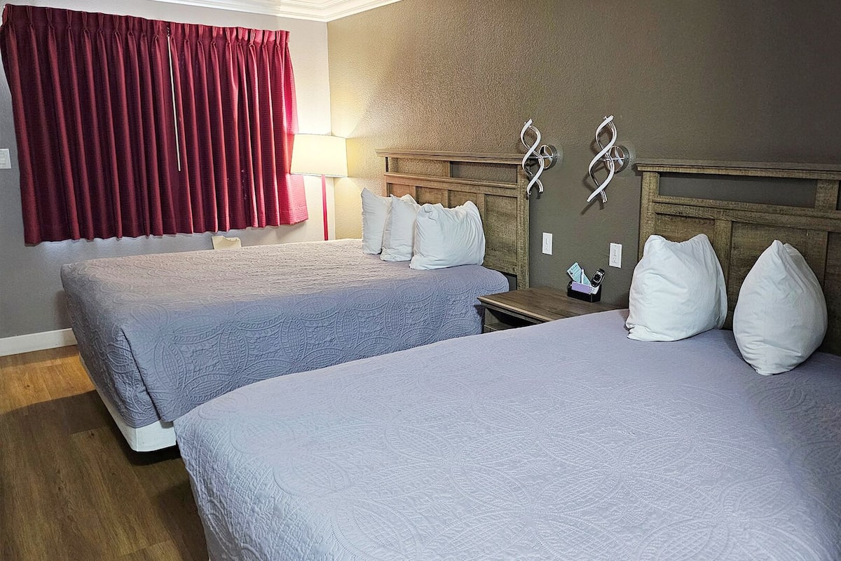 A hotel room with two beds, a wooden headboard, a nightstand, a lamp, and a red curtain.