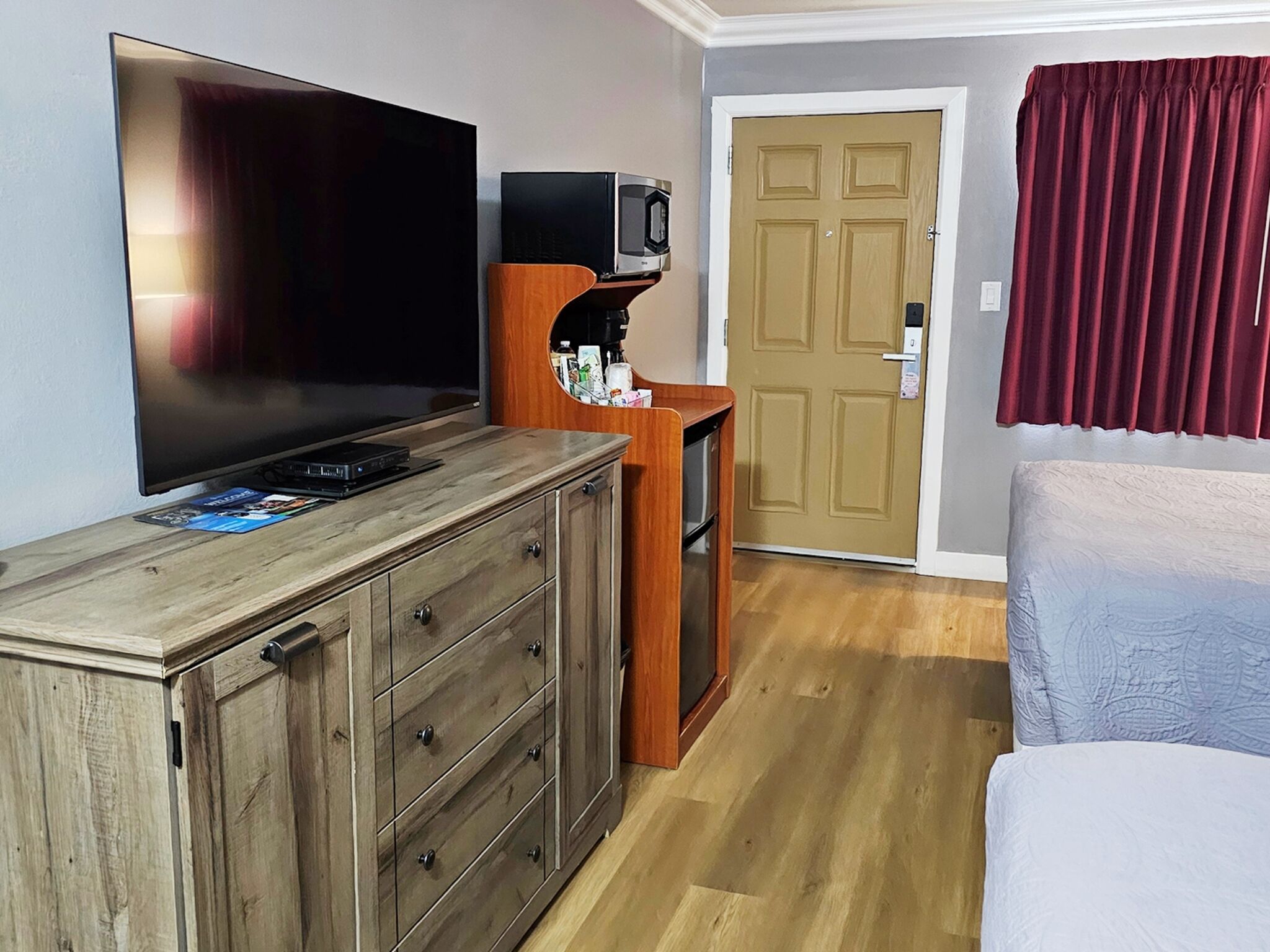 A hotel room with a large flat-screen TV, a wooden dresser, a small wooden shelf with a microwave, a door, and a bed with a red curtain.