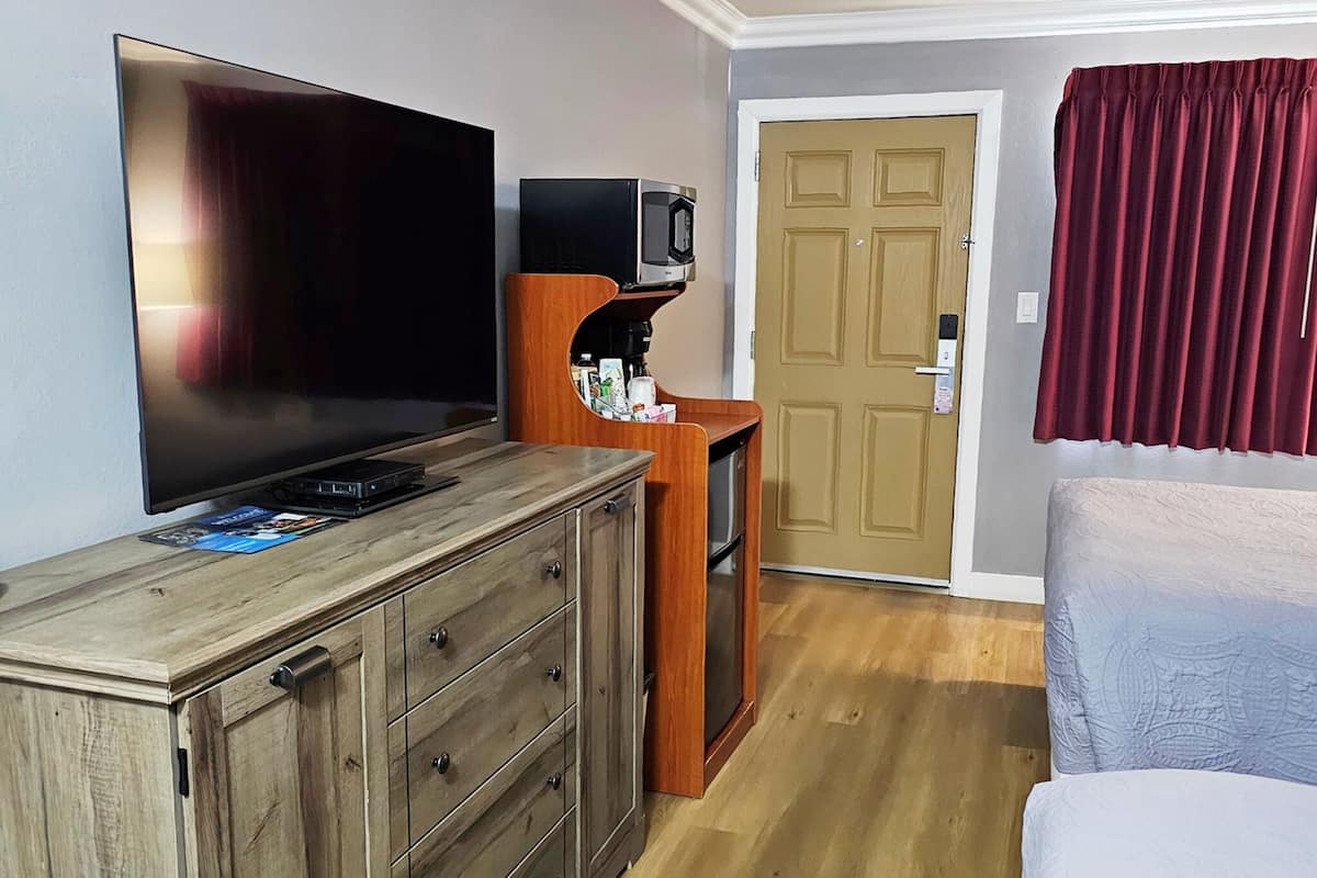 A hotel room with a large flat-screen TV, a wooden dresser, a small wooden shelf with a microwave, a door, and a bed with a red curtain.