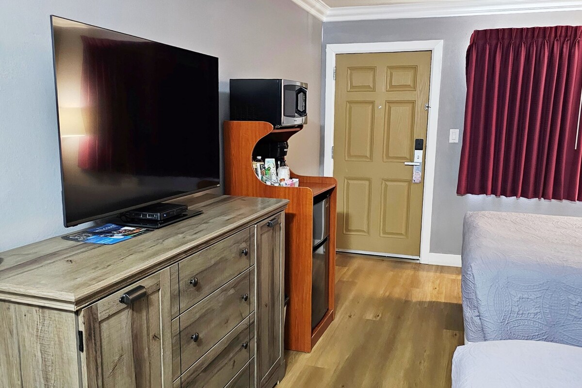 Une chambre d’hôtel dotée d’un grand téléviseur à écran plat, d’une commode en bois, d’une petite étagère en bois avec un four à micro-ondes, d’une porte et d’un lit avec un rideau rouge.