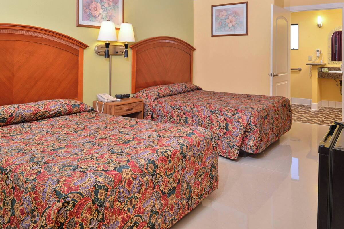 A hotel room with two beds, a wooden headboard, a bedside table with a lamp, a TV, and a bathroom visible through an open door.