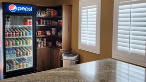 A vending machine stocked with beverages and snacks, a trash bin, and a window with blinds. - Magnuson Hotel Blythe (Blythe)