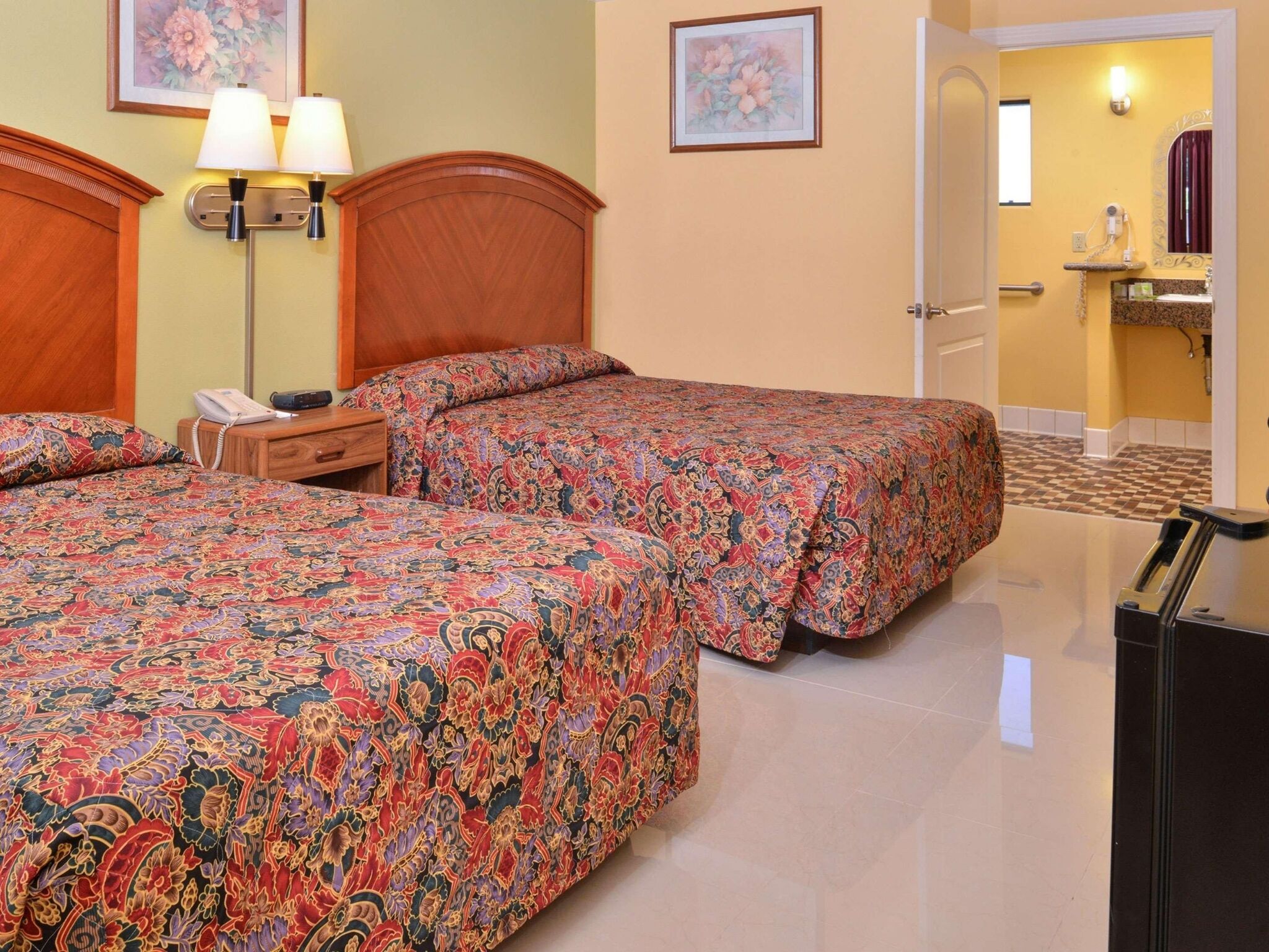 A hotel room with two beds, a nightstand, a lamp, a telephone, and a bathroom visible through an open door.