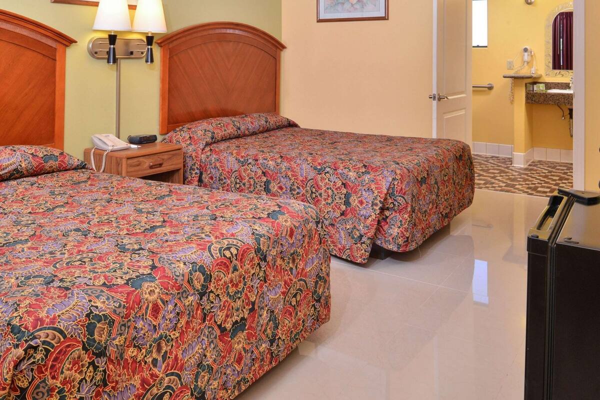 A hotel room with two beds, a nightstand, a lamp, a telephone, and a bathroom visible through an open door.