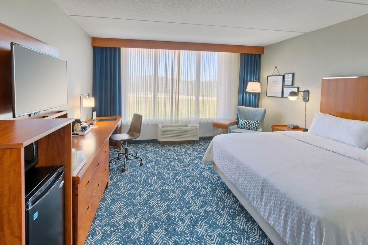A hotel room with a bed, a desk with a flat-screen TV, a chair, a desk lamp, a nightstand, and a window with curtains.