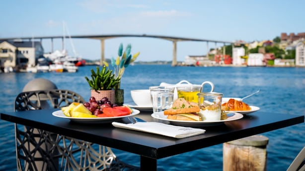 A table with plates of food, a cup, and a small plant by a waterfront with a bridge in the background. - Thon Hotel Kristiansund (Kristiansund)