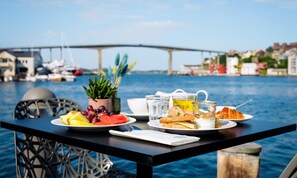 Een tafel met borden eten, een kopje en een kleine plant aan een waterkant, met een brug op de achtergrond.