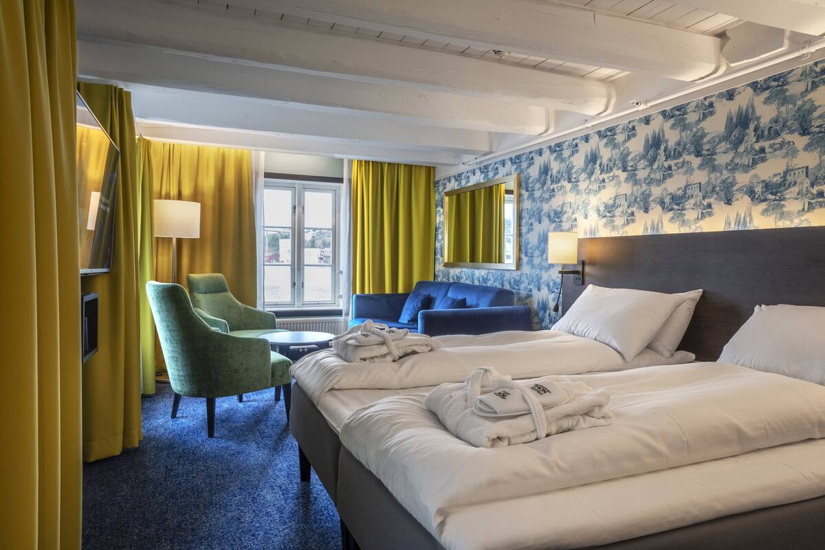 Ein Hotelzimmer mit zwei Betten, einem grünen Sessel, einem kleinen Tisch und einem Fenster mit Vorhängen.