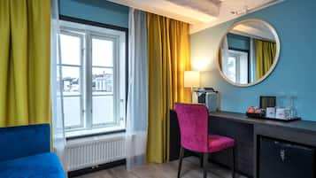A hotel room with a bed, a desk with a chair, a mirror, a window with curtains, and a radiator.