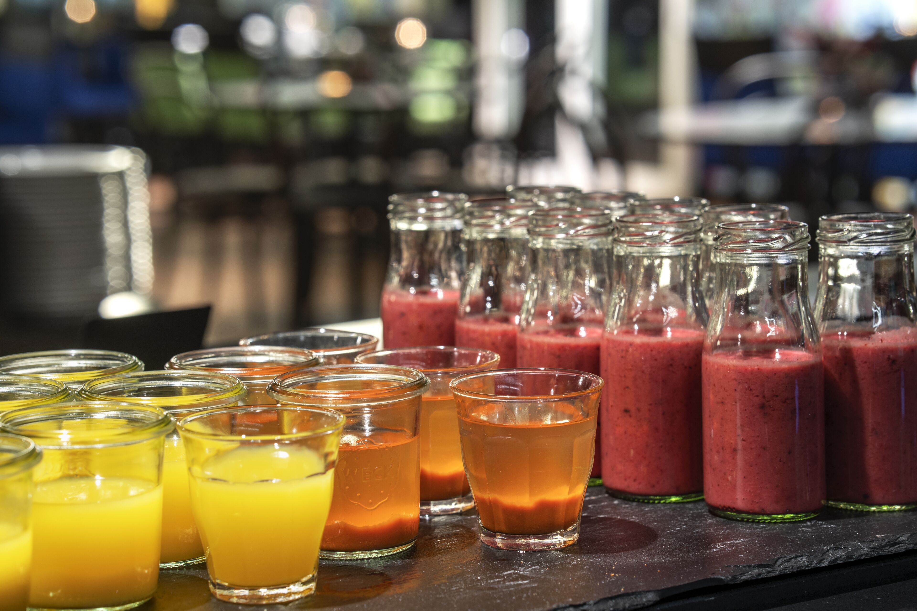 Une variété de boissons, servies dans des verres et des bouteilles, notamment du jus d’orange et des smoothies rouges, sont exposées sur le comptoir du bar.