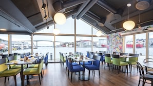 A restaurant with large windows offering views of a bridge and buildings across the water. Tables are arranged with chairs, and the interior has wooden flooring and hanging lights. - Thon Hotel Kristiansund (Kristiansund)