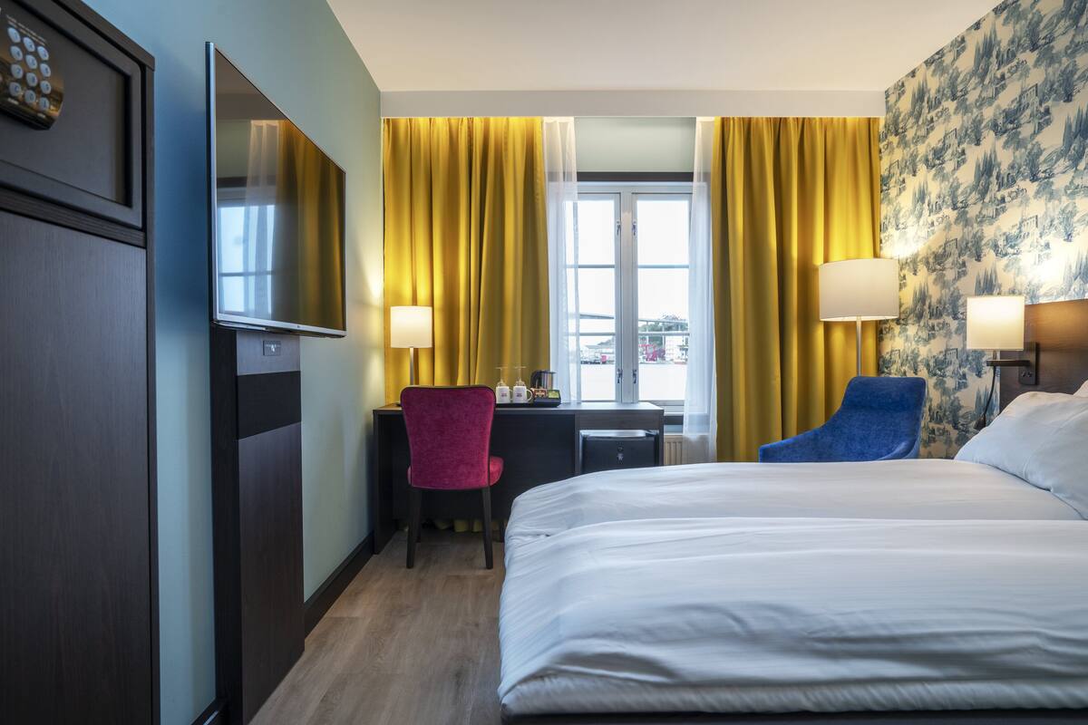 Ein Hotelzimmer mit Bett, Schreibtisch, zwei Stühlen, Fernseher und einem Fenster mit Vorhängen.