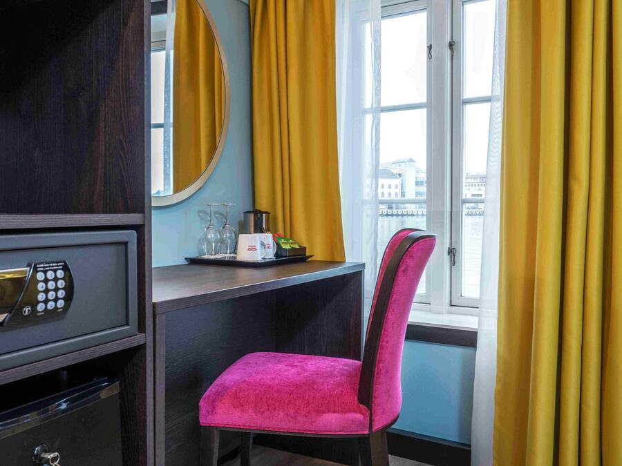 Une chambre d’hôtel équipée d’un bureau, d’une chaise, d’un coffre-fort, d’un miroir et d’une fenêtre avec des rideaux.