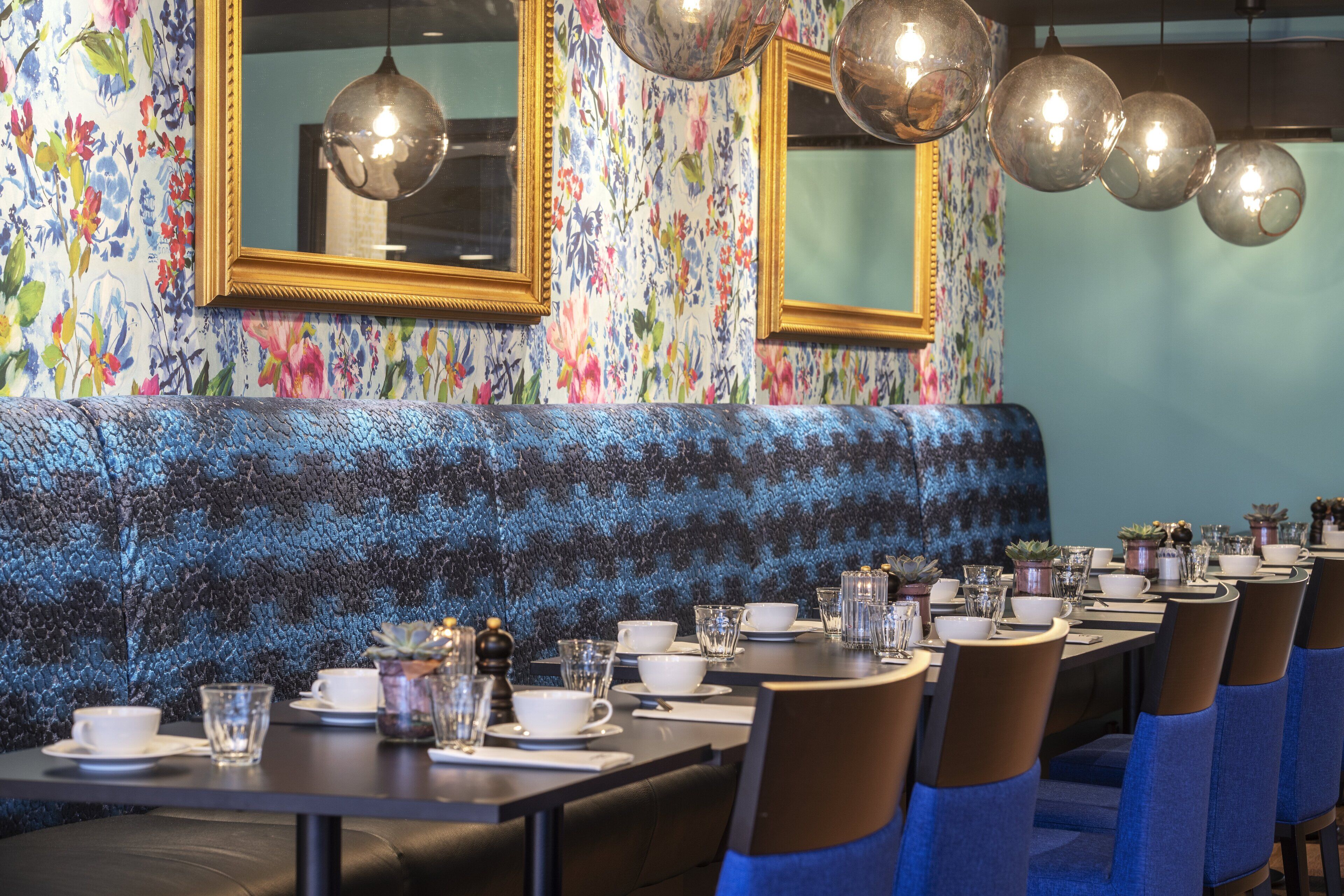 A restaurant with a long table set for dining, a blue upholstered bench, and floral wallpaper.