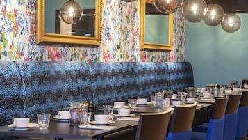A restaurant with a long table set for dining, a blue upholstered bench, and floral wallpaper.