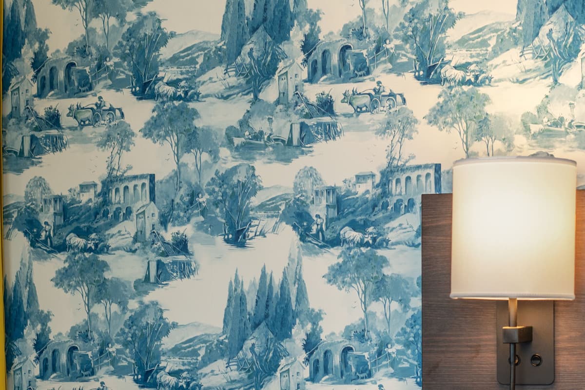 Une pièce dont le papier peint présente un motif bleu et blanc représentant des sc ènes de la campagne, avec une lampe murale en bois et un mur d’accent jaune.