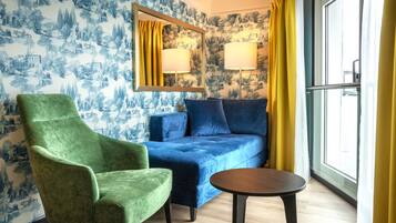 A room with a green armchair, a blue sofa, and a small round table. There is a window with yellow curtains and a mirror with two lamps.