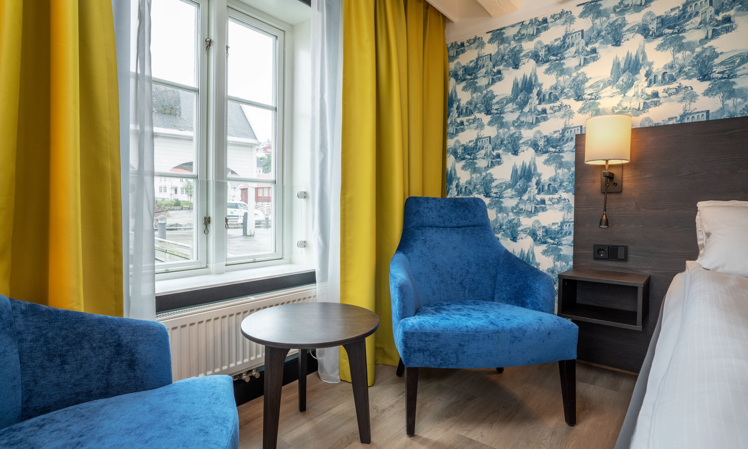 A hotel room with a blue armchair, a small round table, a bed, and a window with yellow curtains.