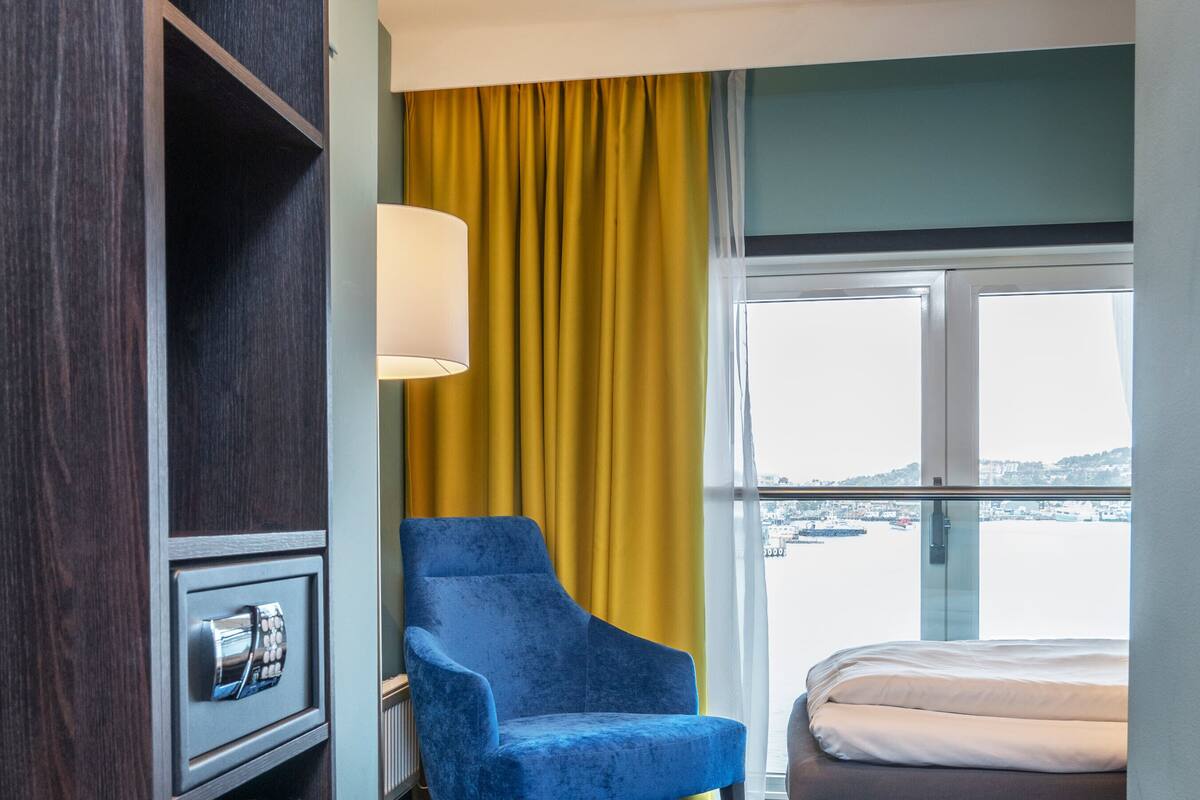 Ein Hotelzimmer mit einem großen Fenster, einem blauen Sessel, einem Bett und einer Minibar.