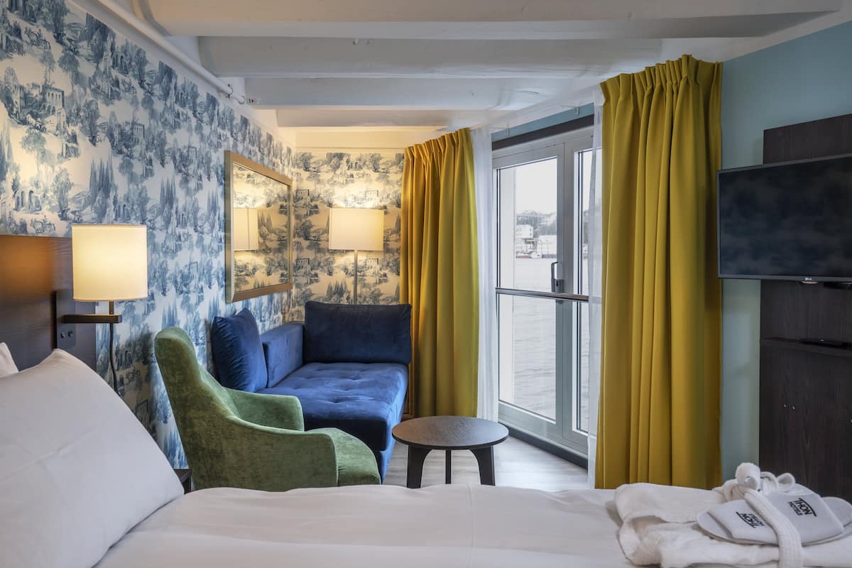 Une chambre d’hôtel avec un lit, un fauteuil vert, un canapé bleu, une petite table ronde, un téléviseur et une fenêtre avec des rideaux jaunes.