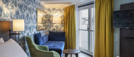 A hotel room with a bed, a green armchair, a blue sofa, a small round table, a TV, and a window with yellow curtains.