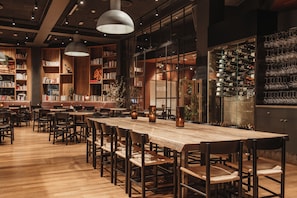 A restaurant with a long wooden table, multiple chairs, hanging pendant lights, and a wine rack with glasses.
