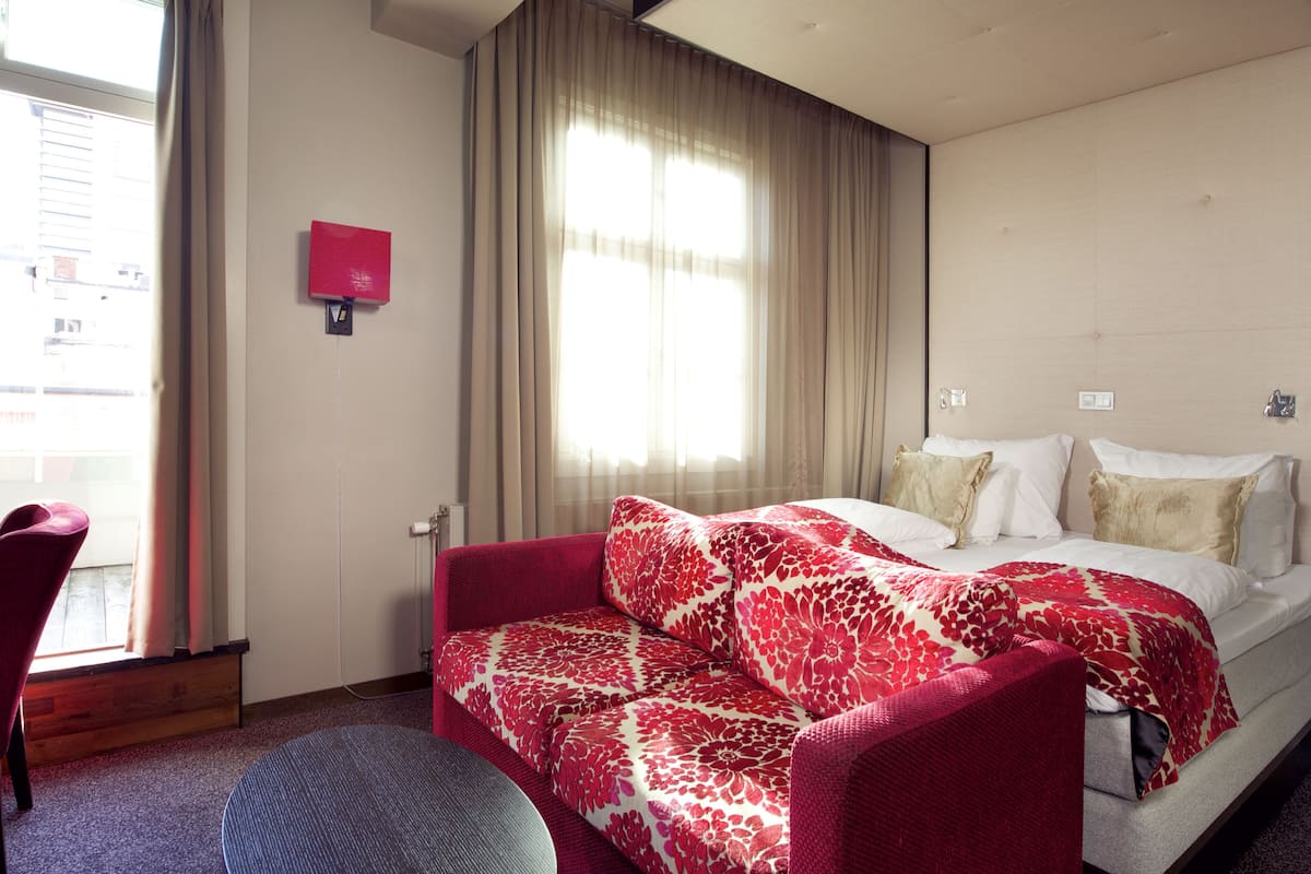 A hotel room with a bed, a red sofa, a small table, and a window with curtains.