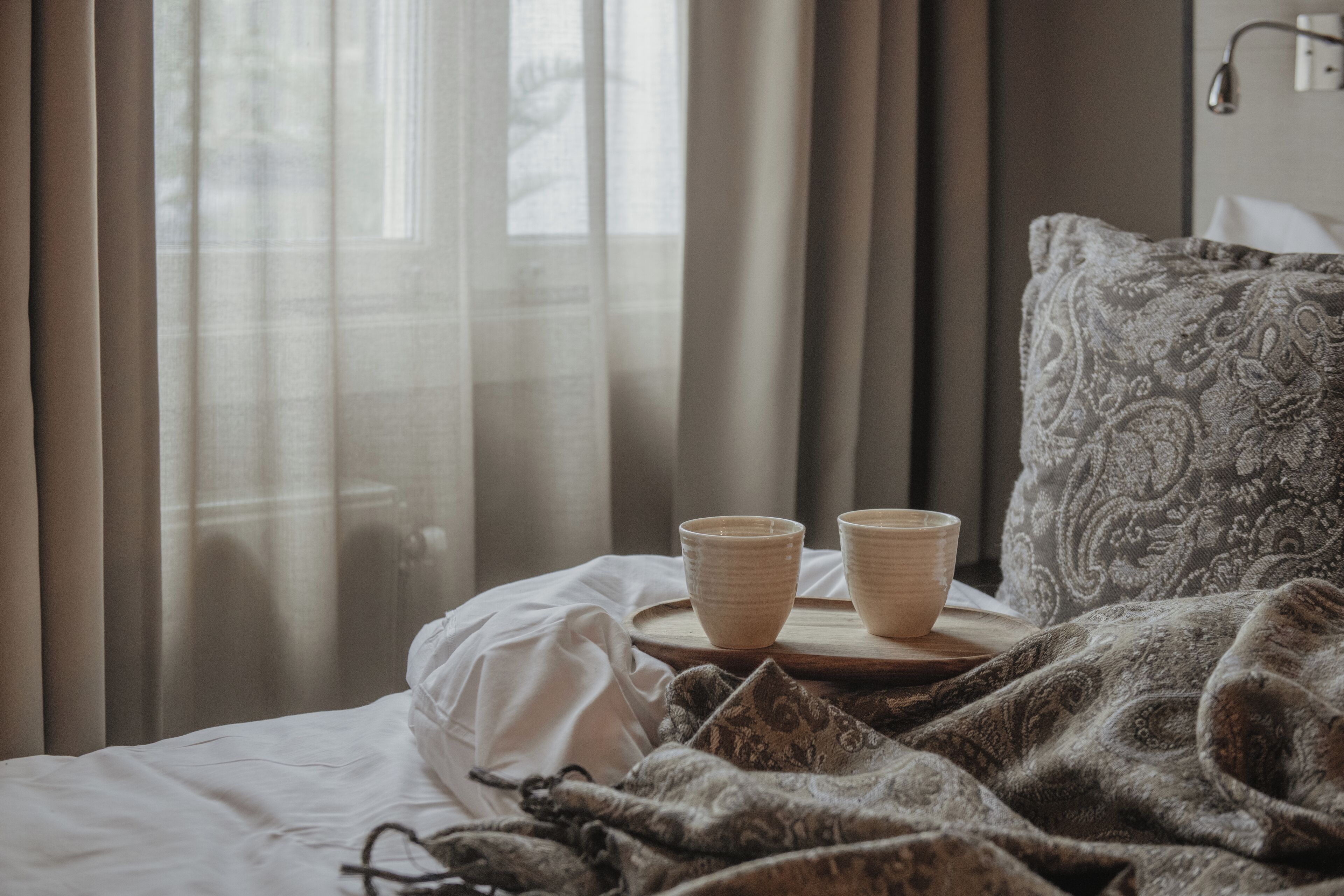Ein Bett mit weißer Decke, einer gemusterten Wolldecke und zwei beigen Tassen auf einem Holztablett.