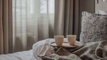 Ein Bett mit weißer Decke, einer gemusterten Wolldecke und zwei beigen Tassen auf einem Holztablett.