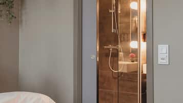 A modern bathroom with a glass shower enclosure, a bed with a white cover, and a wall-mounted light switch.