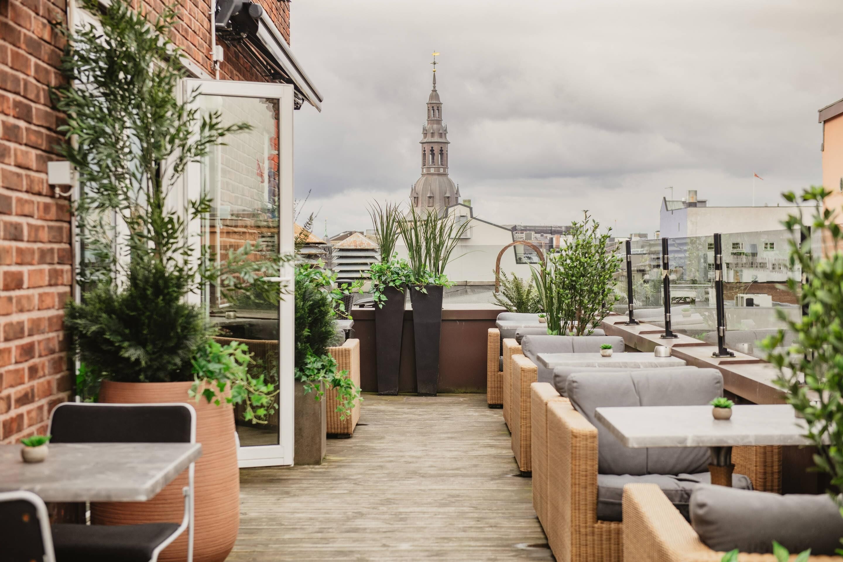 Une terrasse sur le toit, aménagée avec des meubles en osier, des plantes en pot et offrant une vue sur le clocher d’une église.