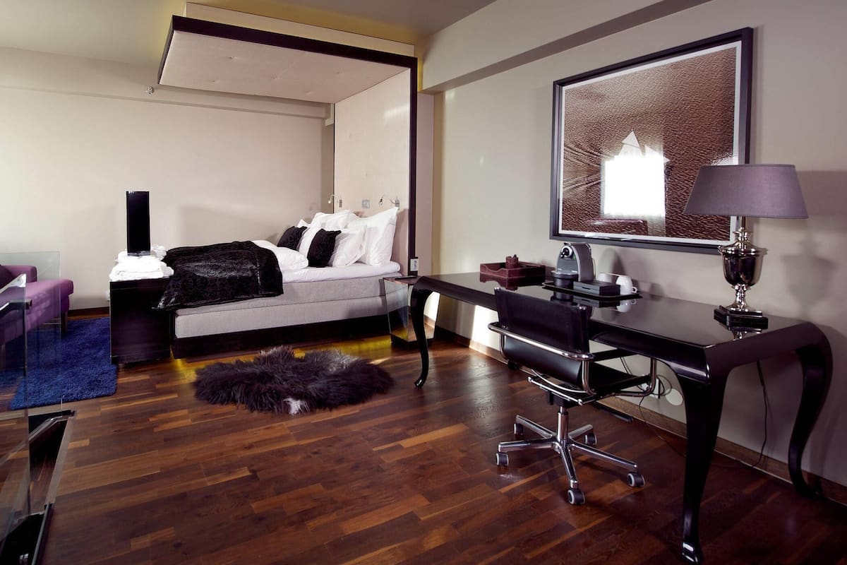 A modern hotel room with a bed, a desk, a chair, a sofa, and a glass table.