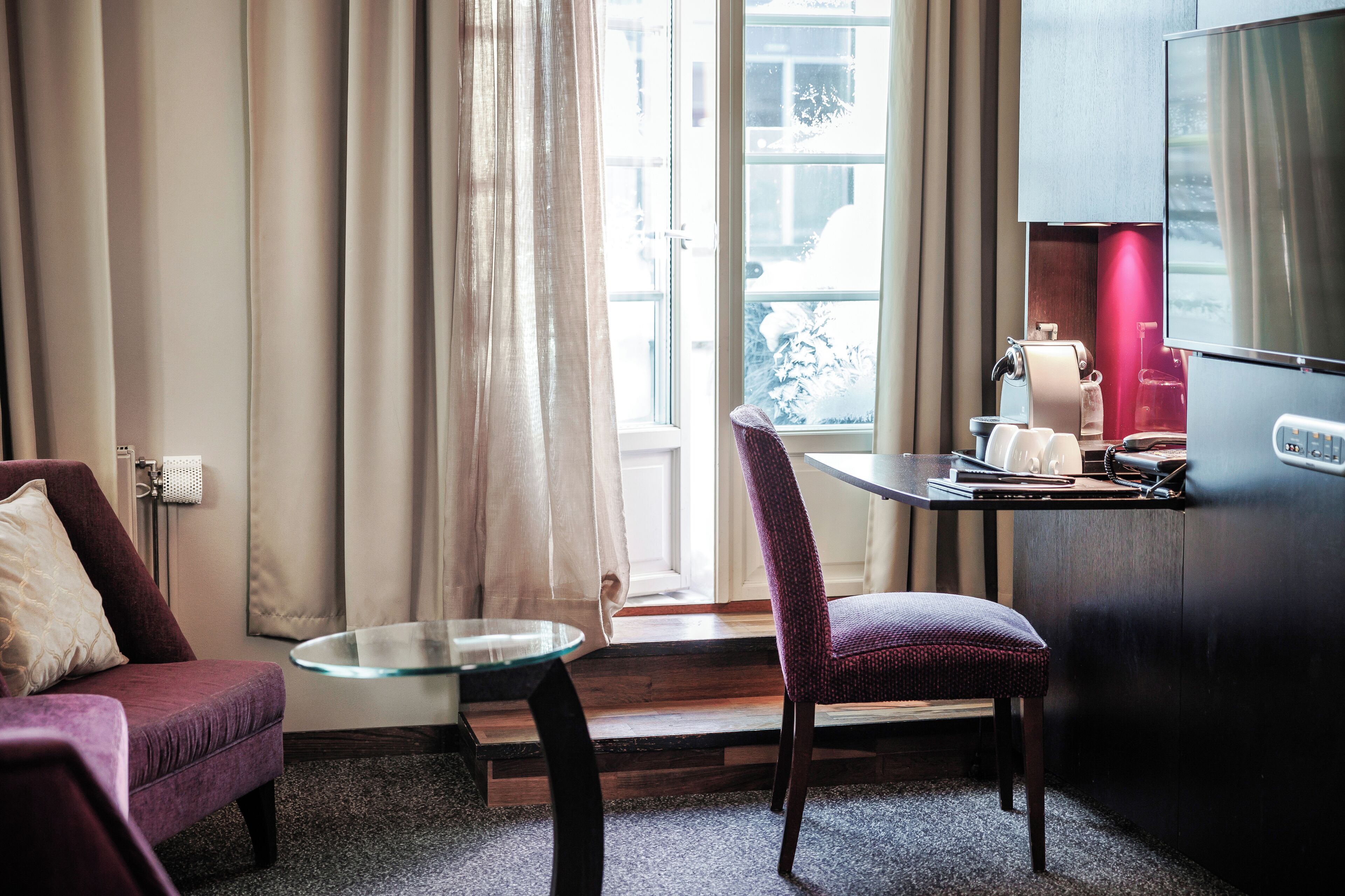 A modern hotel room with a glass table, a purple chair, a sofa, and a view of the outdoors through a large window.