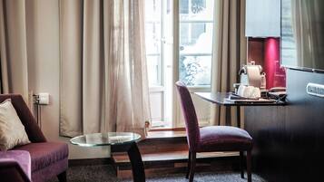A modern hotel room with a glass table, a purple chair, a sofa, and a view of the outdoors through a large window.