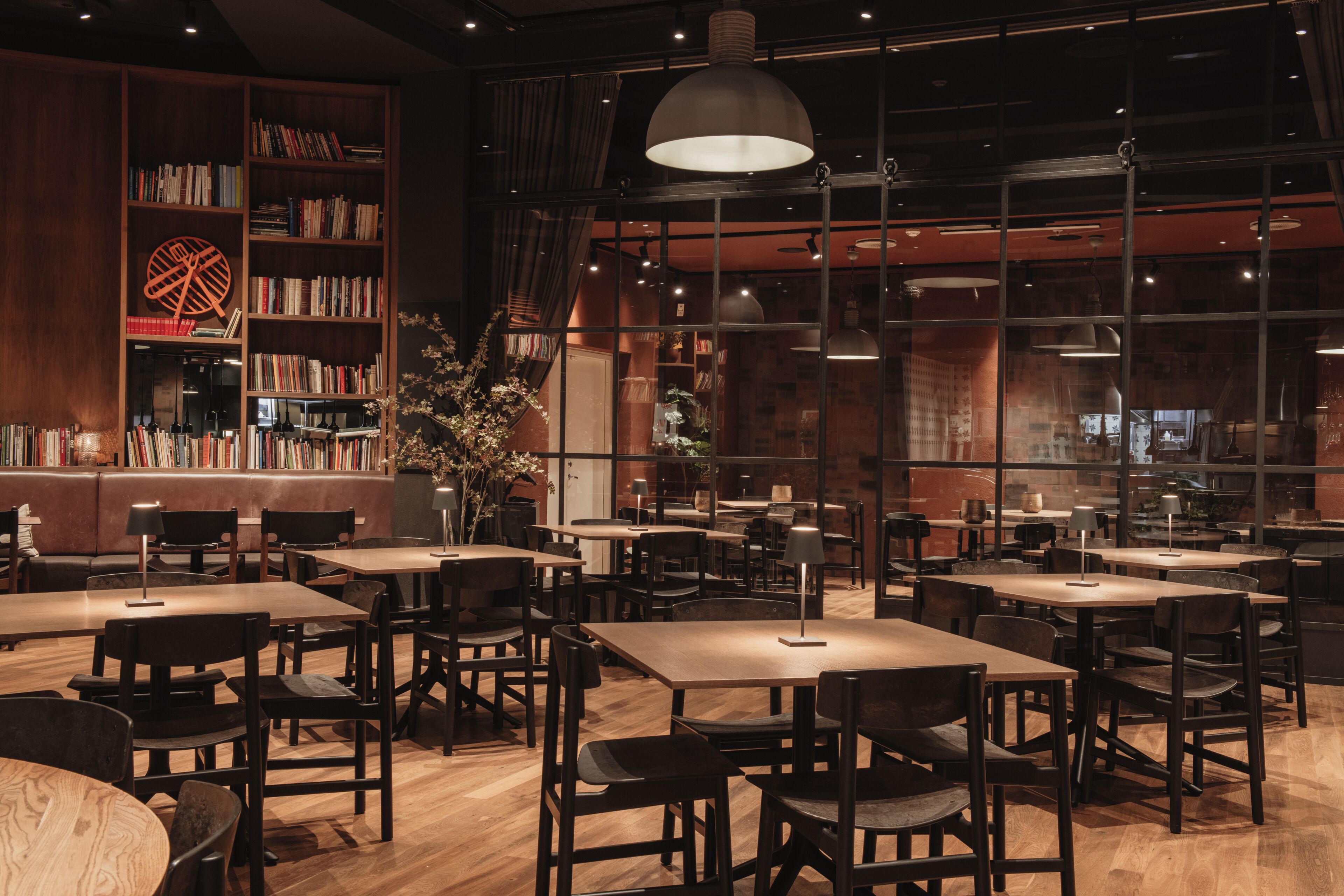 A restaurant with wooden tables and chairs, a bookshelf, and large windows.