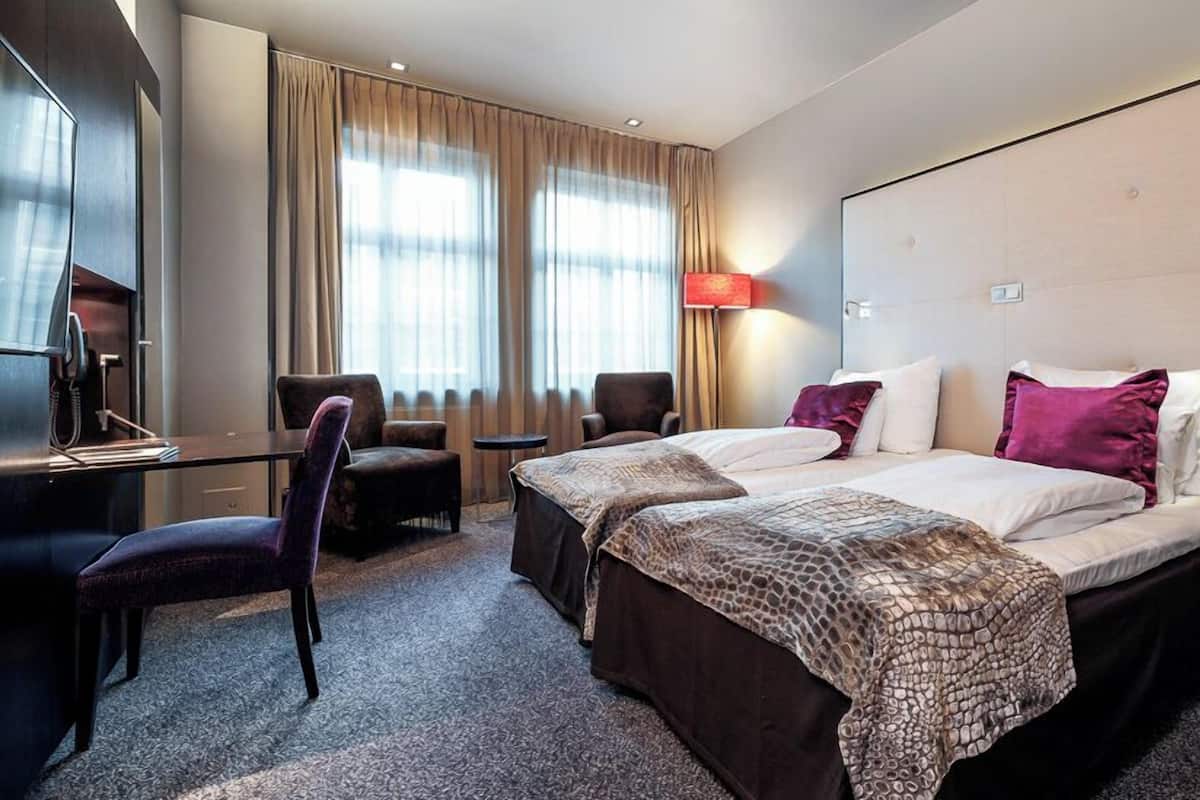 A hotel room with a large bed, two bedside tables, a desk, a chair, and a large window with curtains.