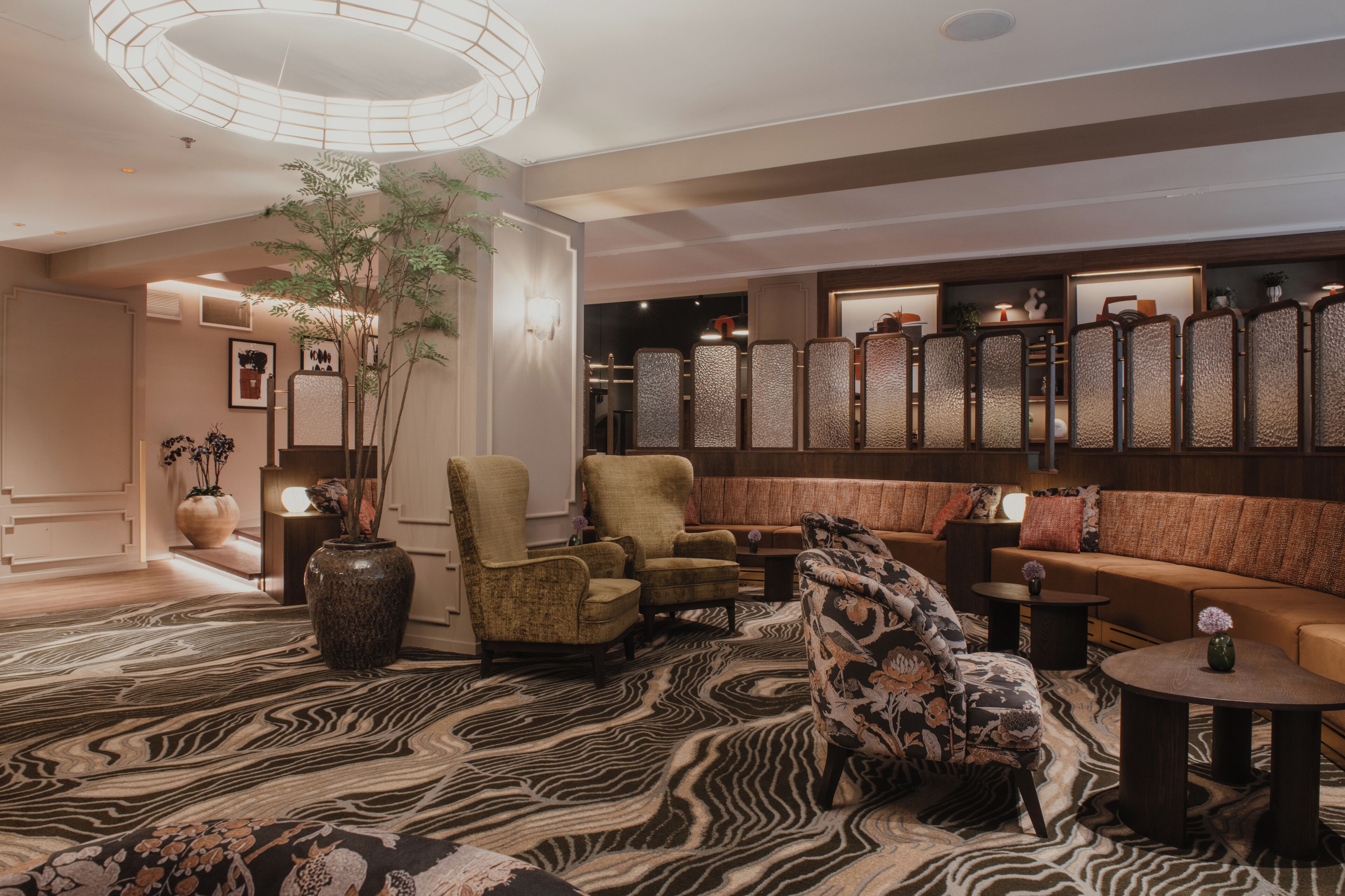 Un moderno vestíbulo de hotel con un sofá, sillones, un tapete estampado, un jarrón grande y un panel decorativo en la pared.