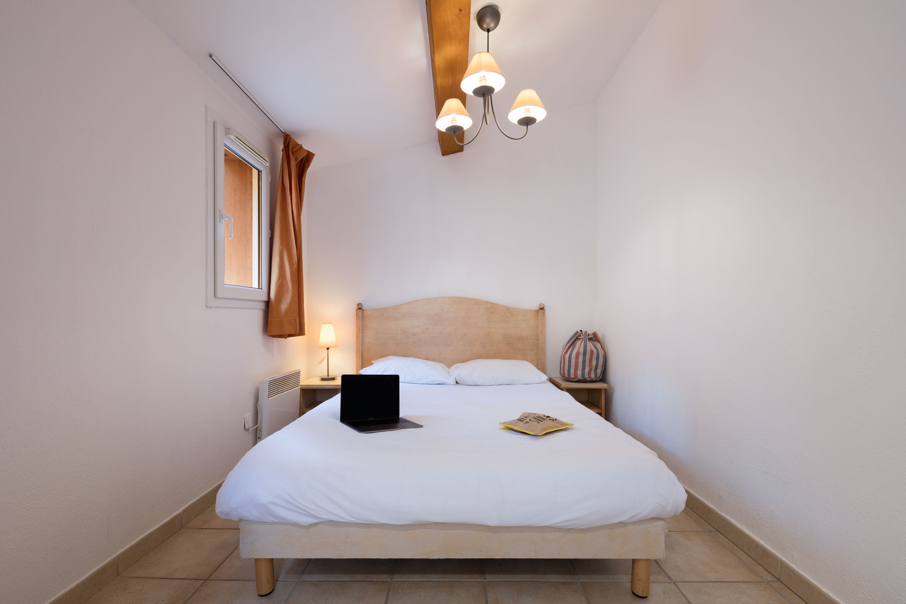 A neatly made bed with a laptop and a book, a bedside lamp, and a window with curtains.