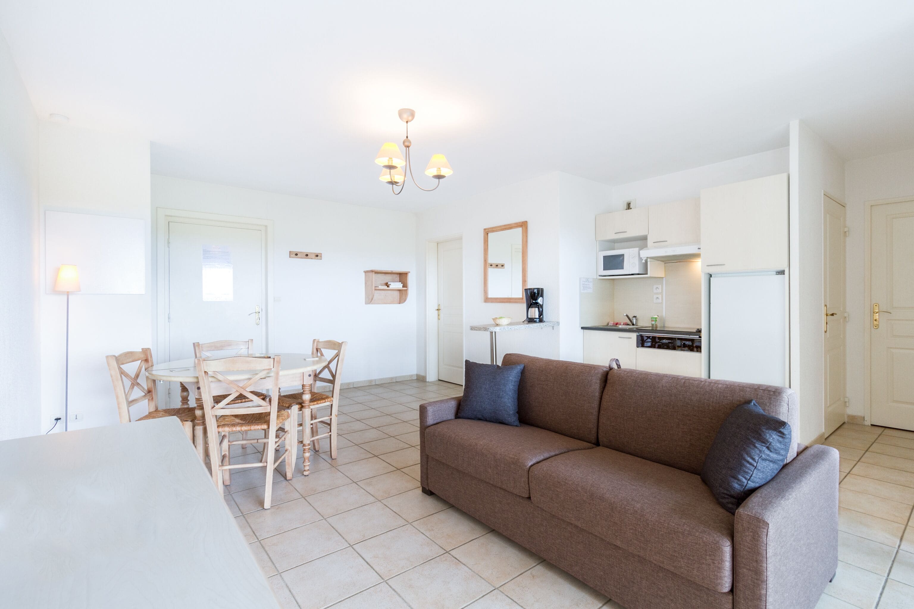 A modern living room with a brown sofa, wooden dining table, and chairs. A kitchen area is visible in the background with white cabinets and appliances.