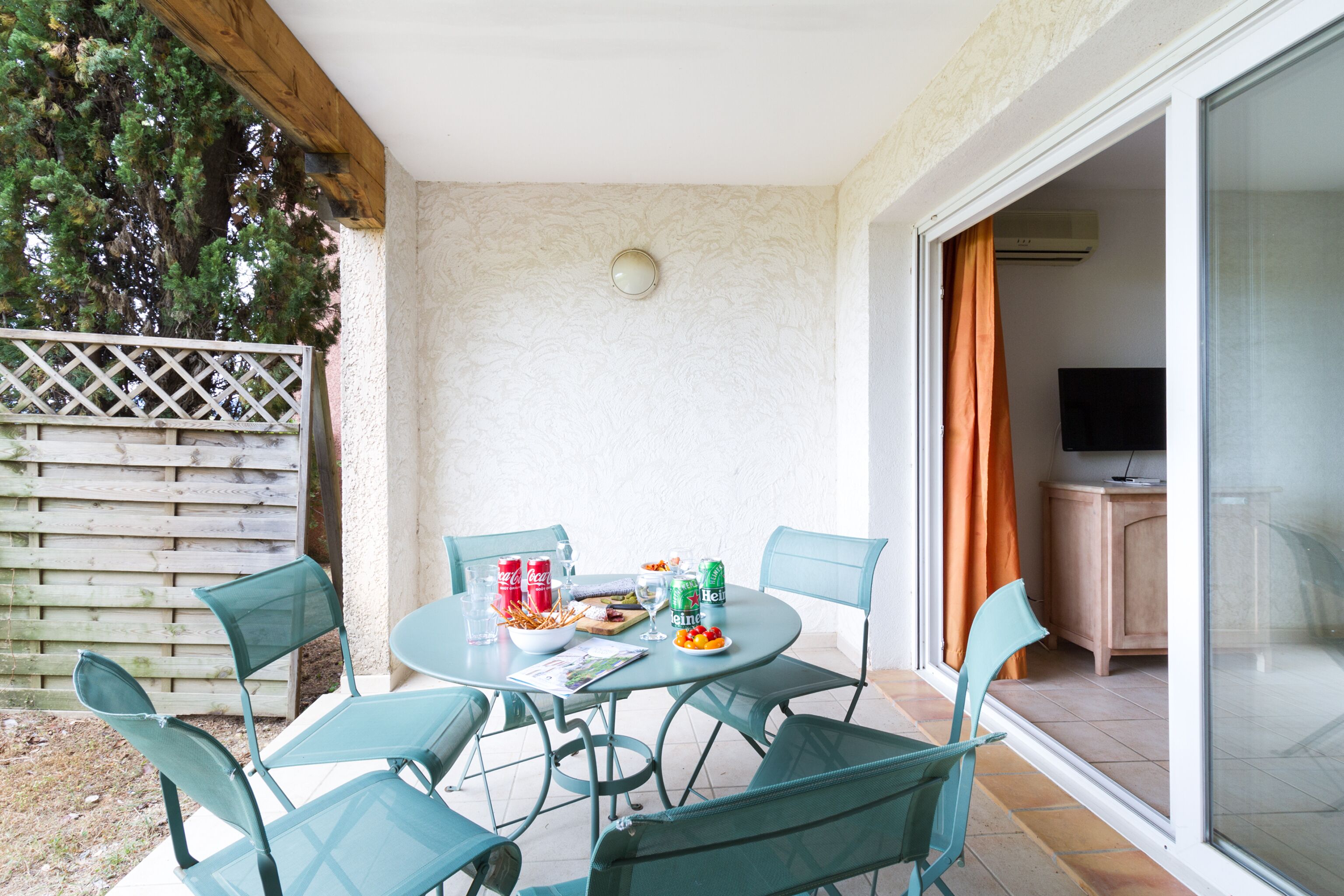 En terrasse med et rundt bord og fire stole, et træhegn og en skydedør, der fører til et rum med et fjernsyn.