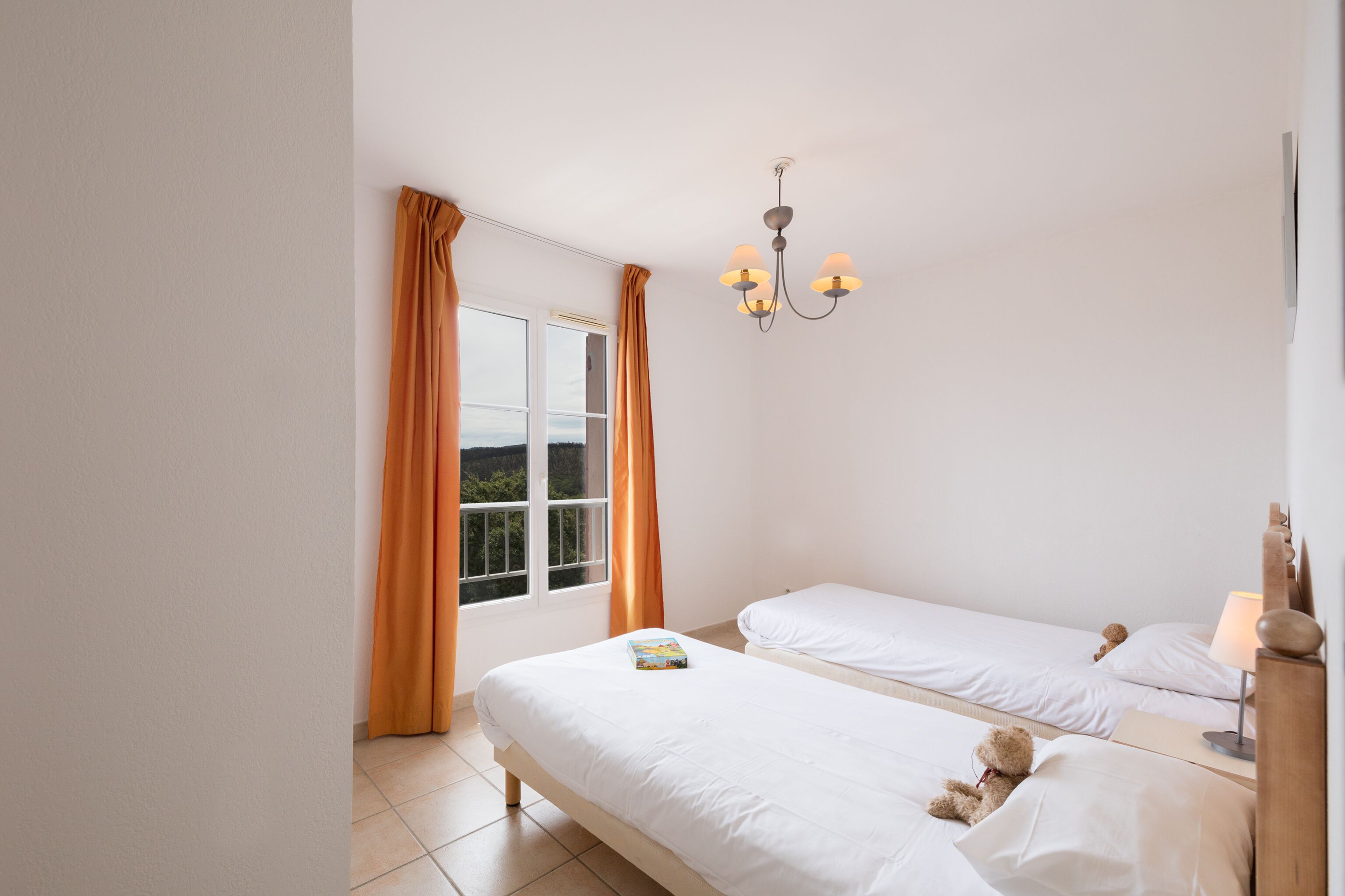 A bedroom with two beds, a window with curtains, a chandelier, and a bedside table with a lamp.