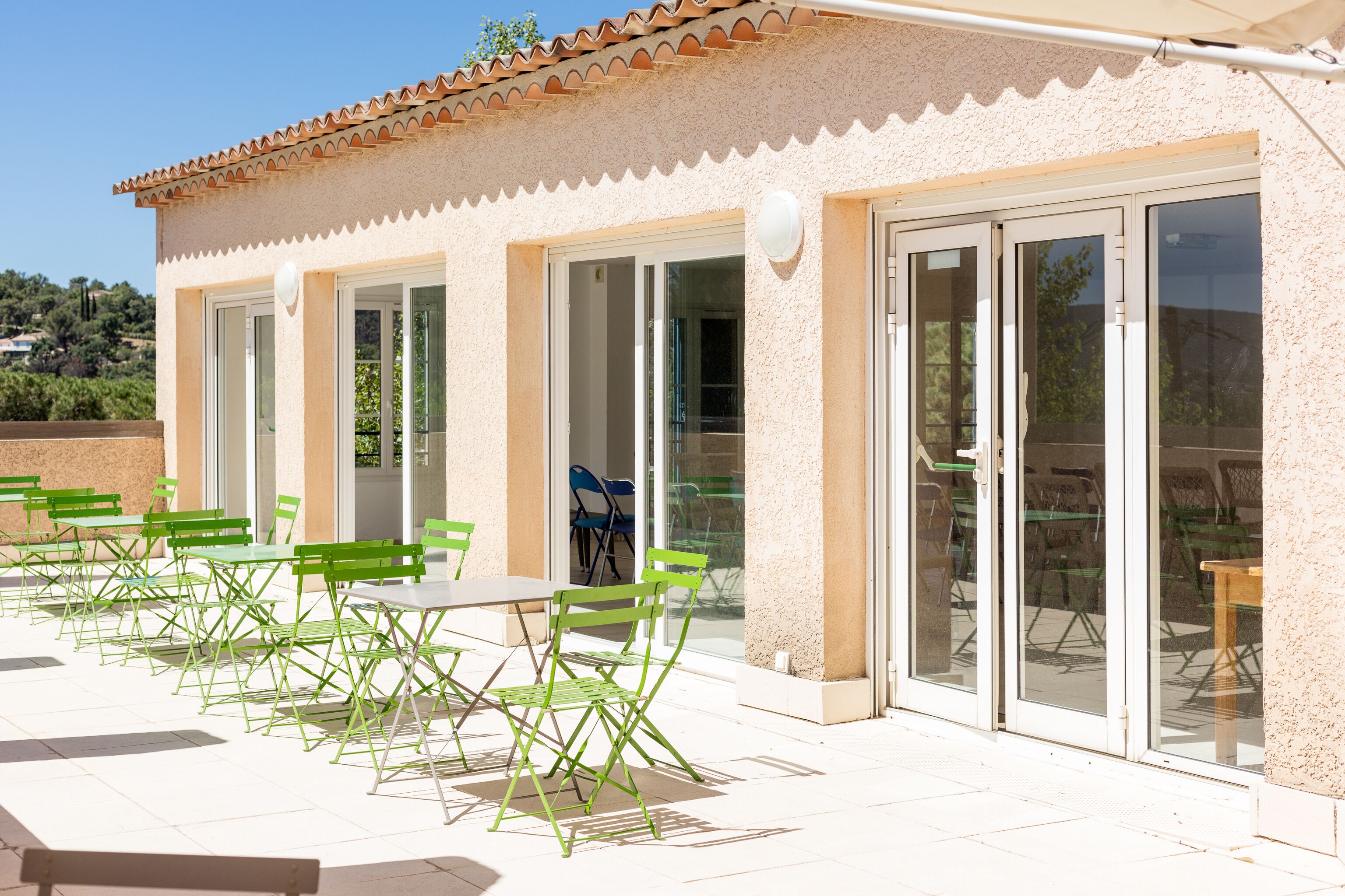 An outdoor patio with green chairs and tables, a building with a tiled roof, and glass doors leading to an interior space.