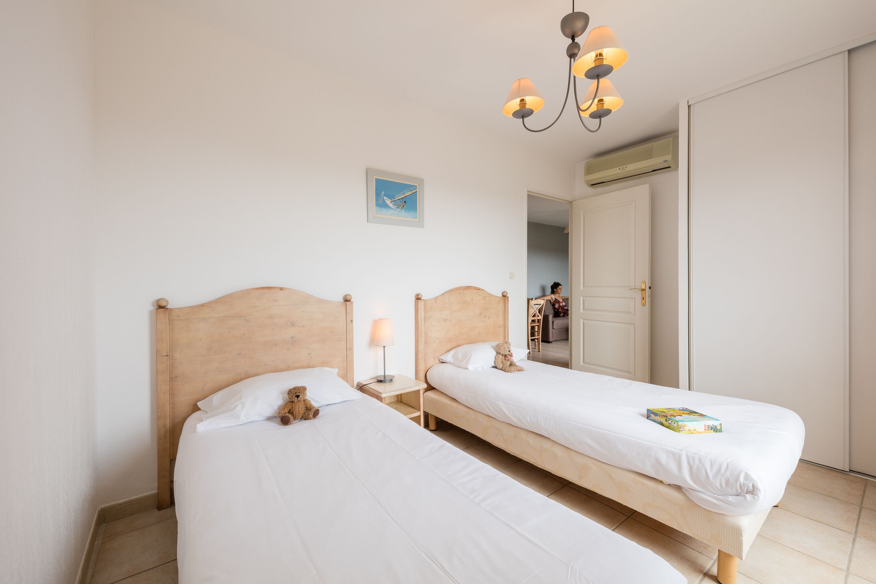 Une chambre d’hôtel avec deux lits simples, une tête de lit en bois, une table de chevet avec une lampe, un luminaire au plafond et un tableau encadré au mur.