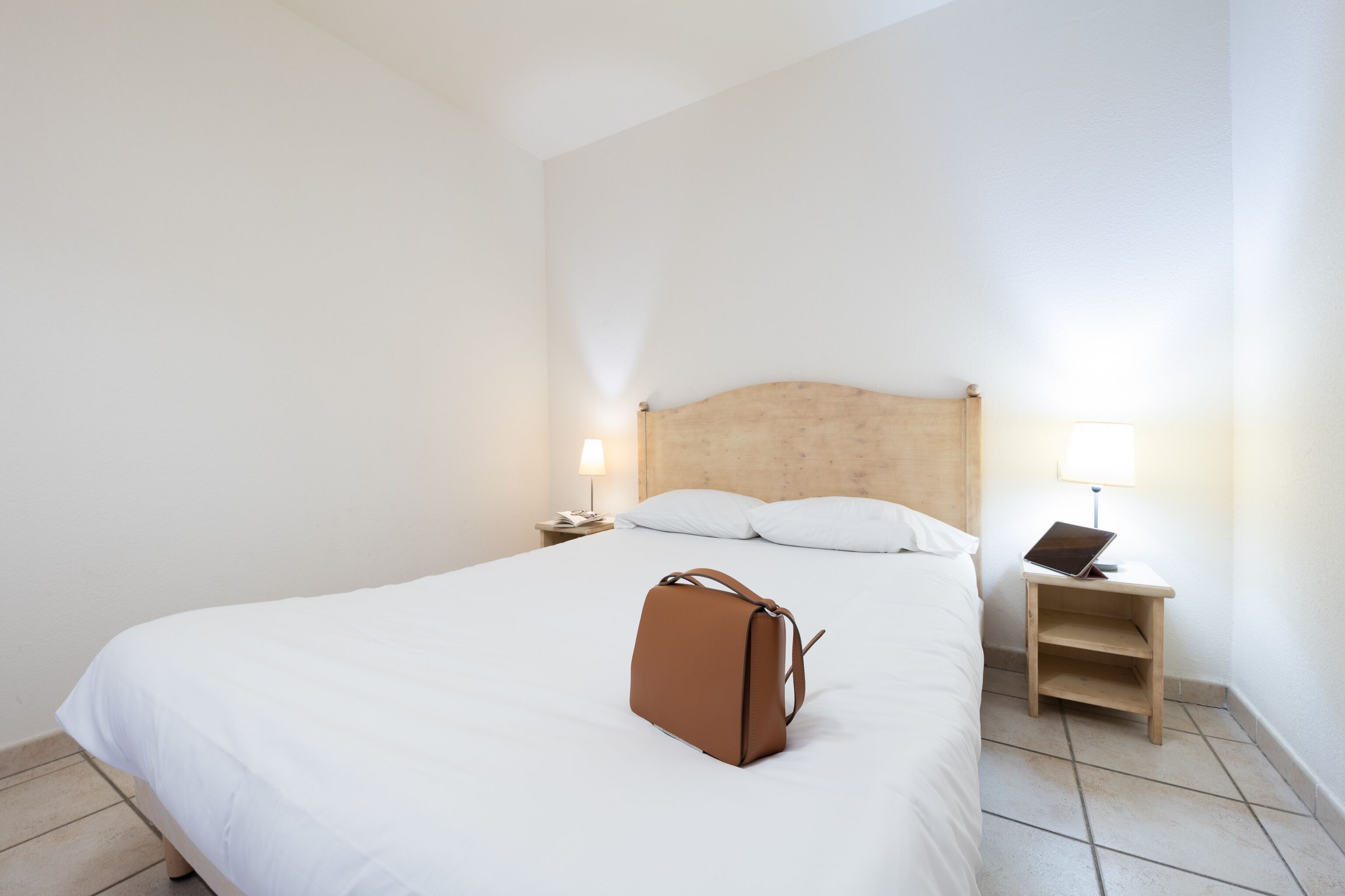 A neatly made bed with white linens, a wooden headboard, and a brown handbag placed on the bed. There is a bedside table with a lamp and a phone.