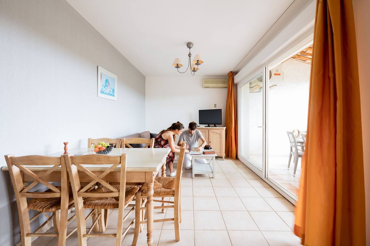 Un coin repas avec des meubles en bois, un téléviseur et une vue sur l’extérieur, visible à travers une porte vitrée.