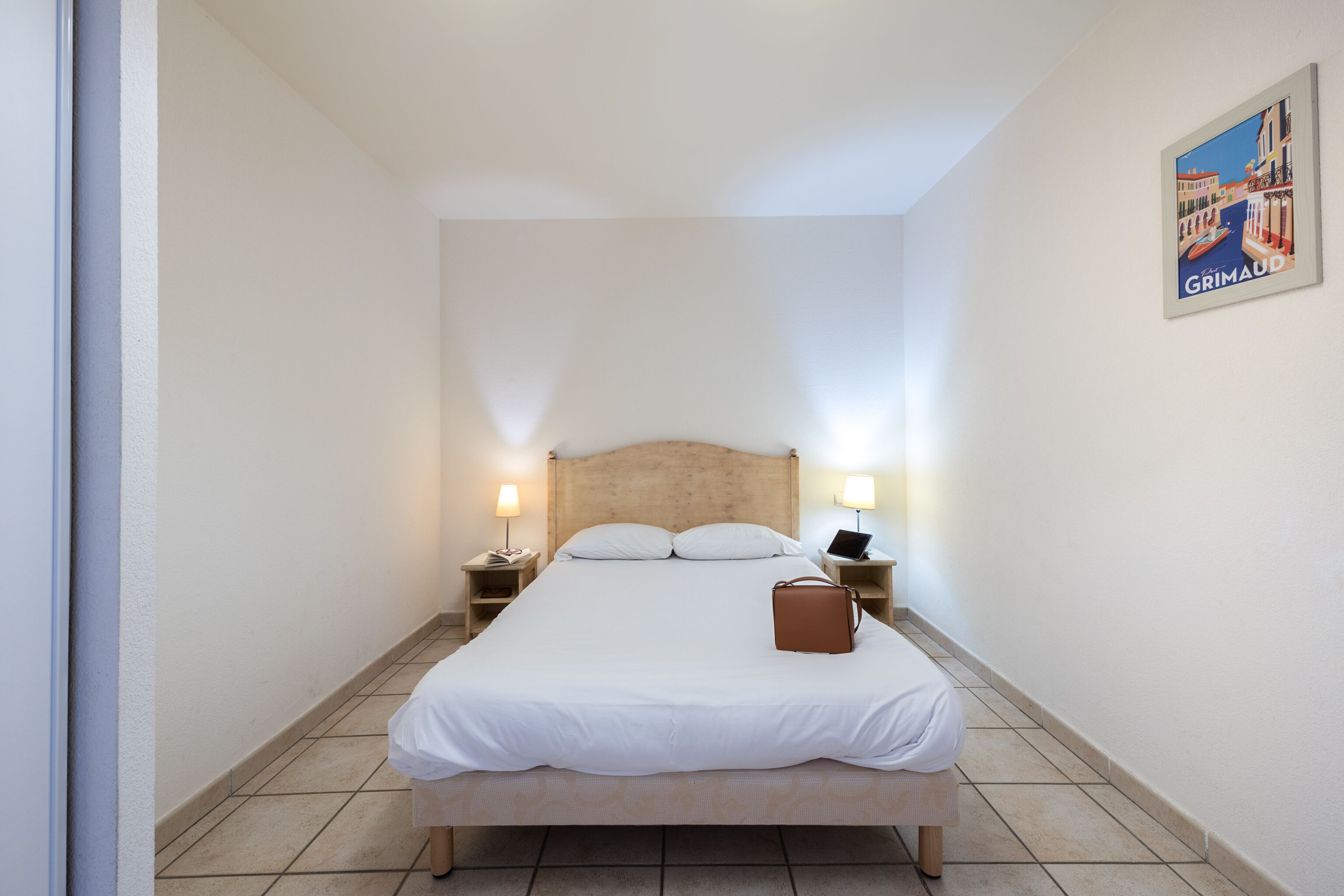A neatly made bed with white linens, two bedside lamps, a laptop on one nightstand, and a brown handbag on the bed.
