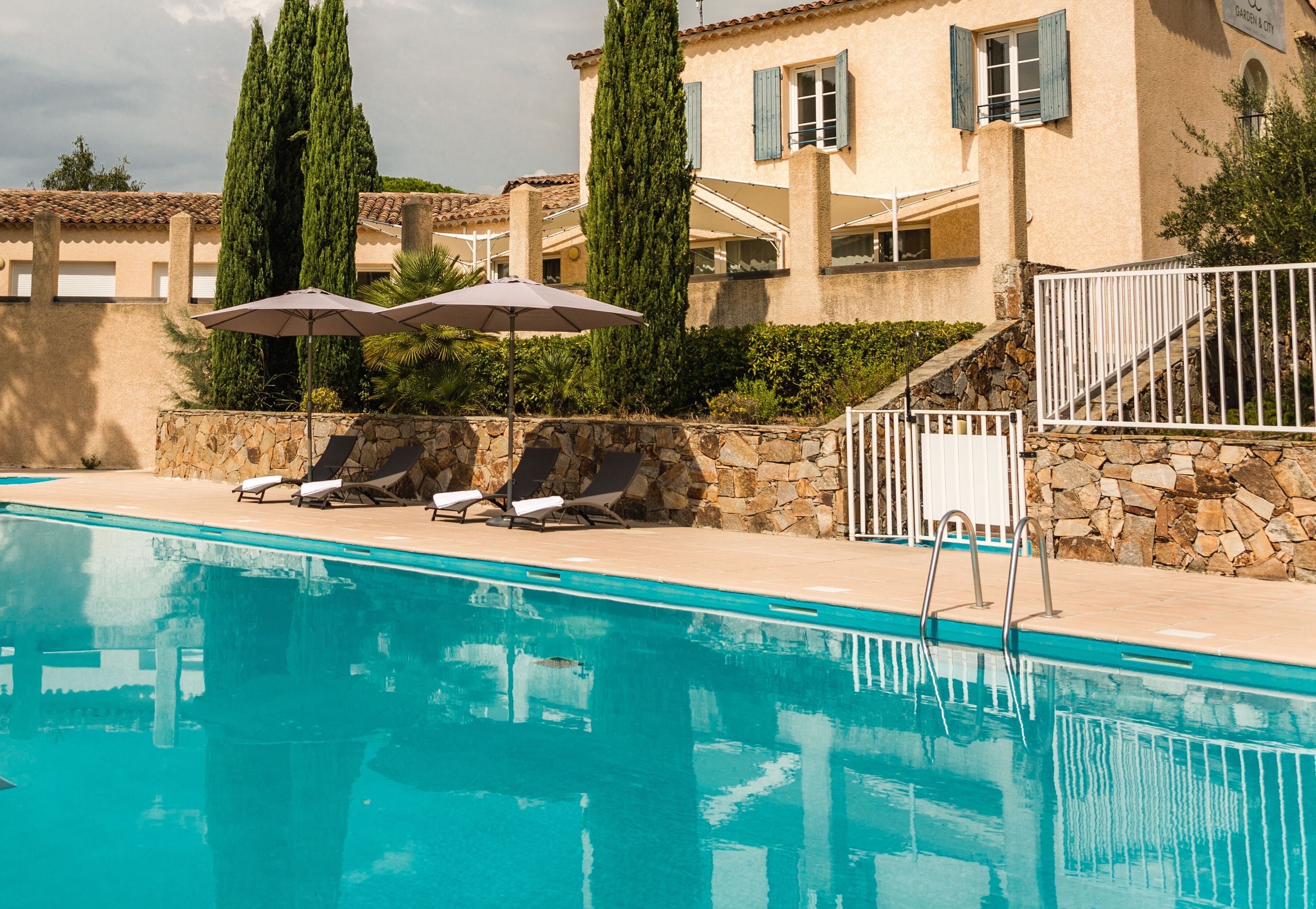 A swimming pool with lounge chairs and umbrellas, a stone wall, and a building with a tiled roof and shutters.