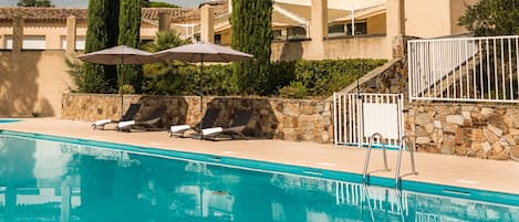 A swimming pool with lounge chairs and umbrellas, a stone wall, and a building with a tiled roof and shutters.