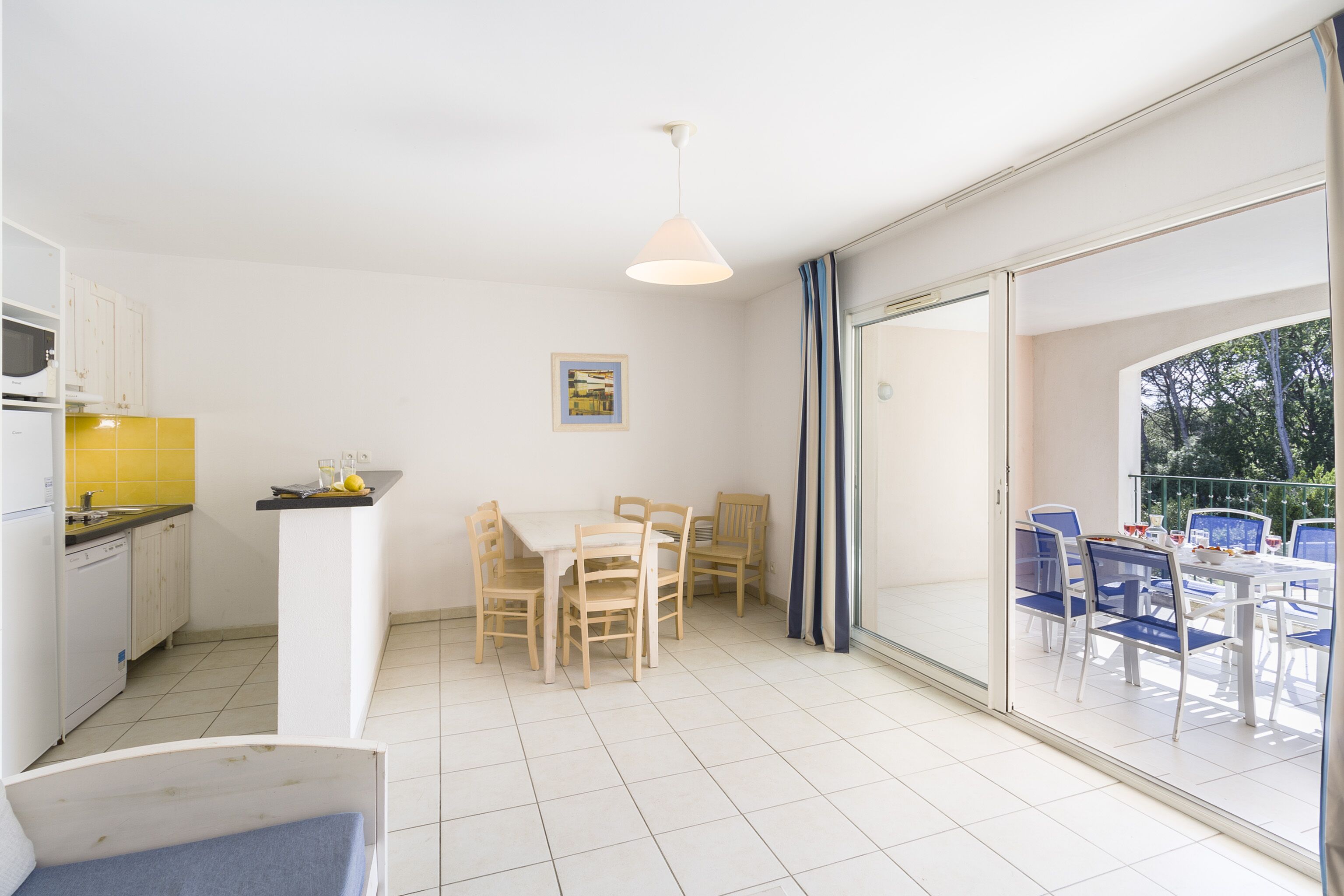 Una cocina moderna con armarios blancos y un salpicadero amarillo, una zona de comedor con sillas de madera y una mesa blanca, y un balcón con sillas azules y un ventanal de vidrio.