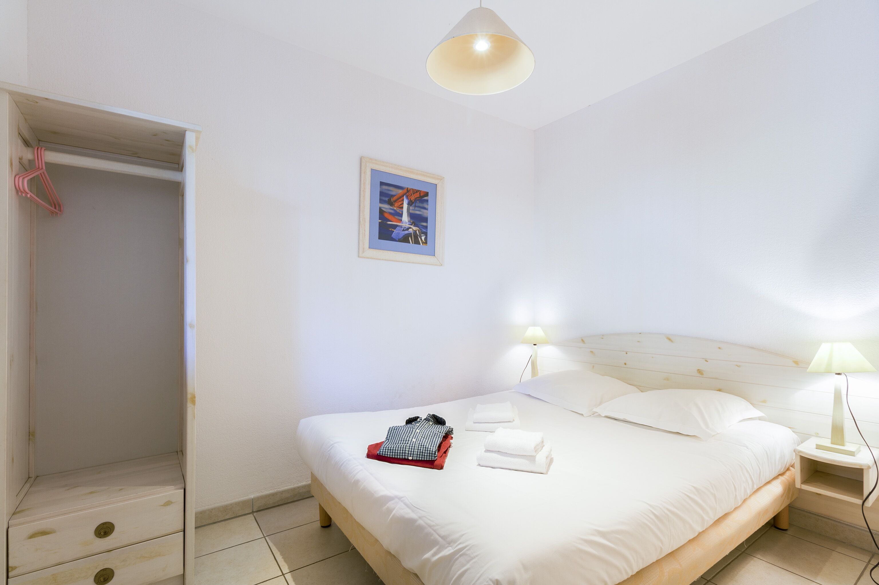 Un lit bien fait, avec des oreillers et des serviettes pliées, une lampe de chevet et un tableau encadré au mur.